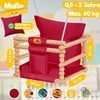 A red baby swing made of wood and fabric, with measurement indicators and text overlays. Produktbild-Vorschau