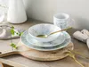 A set of dishes with a marble-like pattern, including plates, a bowl, and a mug, arranged on a wooden board with cutlery and a sprig of greenery. Náhľad obrázku produktu