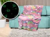 A fluffy blanket with a unicorn and rainbow pattern is draped over a teal sofa. A circular inset shows a close-up of the blanket's glow-in-the-dark unicorn pattern on a black background. Anteprima dell’immagine prodotto