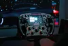A racing steering wheel with a digital display and various buttons and knobs, set against a blurred background of a car interior and starry sky. Produktbild-Vorschau