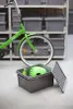 A green bicycle is parked in front of shelves filled with gray storage boxes. In the foreground, an open gray storage box contains a green helmet and a green bottle. Anteprima dell’immagine prodotto