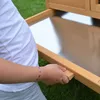 A person's arm and hand are shown pulling out a metal tray from a wooden structure, possibly a pet house or hutch. Produktbild-Vorschau