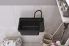 A black sink with a black faucet is mounted on a white tiled wall in a laundry room or garage setting. Laundry baskets and cleaning supplies are visible around the sink. Anteprima dell’immagine prodotto