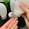 A hand is holding a clear travel bottle and dispensing liquid onto a person's palm. Other travel bottles and a pink cap are visible in the background. Produktbild-Vorschau