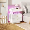 A white nightstand with two drawers and an open storage shelf with LED lighting, placed next to a bed. An easel with a blank canvas is on the left. Podgląd zdjęcia produktu