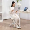 A woman is sitting on a white office chair while reading a magazine. A laptop and lamp are on the desk behind her. Podgląd zdjęcia produktu