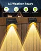 Two solar fence lights mounted on a wooden fence, illuminating downwards with yellow light. The image also shows icons and text indicating weather resistance. Produktbild-Vorschau
