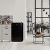 A black mini-fridge is placed in a kitchen-like setting next to cabinets and a patterned backsplash, with a dining area visible through a partition in the background. Voir les images du produit