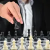 A person's hand is holding a black chess king over a chessboard with black and white pieces. Produktbild-Vorschau