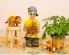 A young boy in sunglasses and a baseball cap holds two plush triceratops dinosaurs. Another plush triceratops is on a wooden stool to the left, and another is on the floor to the right, near a large potted plant. Anteprima dell’immagine prodotto