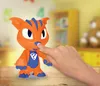 A child's hand is touching the nose of an orange and blue toy creature on a wooden surface. The background is blurred and appears to be a child's room with shelves and a teddy bear. Voir les images du produit