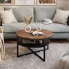 A round coffee table with a wooden top and black metal legs is placed on a fluffy rug in front of a light blue sofa. Decorative items are on the table and sofa. Produktbild-Vorschau