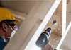A construction worker wearing safety gear is drilling into wooden beams. Produktbild-Vorschau