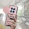 A hand holding a rose gold phone case with a camera cover and ring holder, with a blurred background of an indoor setting. Produktbild-Vorschau