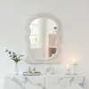 A fluffy white mirror is mounted on a white wall above a marble-topped cabinet. A vase with flowers, books, and decorative objects are on the cabinet, along with two lit candles in brass holders. Produktbild-Vorschau