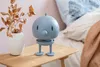 A light blue Hoptimist LED lamp sits on a wooden bedside table next to a vase of dried leaves and a bed with a peach blanket. Produktbild-Vorschau