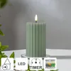 A green LED candle with a flickering flame sits on a clear glass coaster, with greenery in the foreground and a blurred background. Podgląd zdjęcia produktu