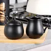 A black sugar bowl with a spoon and a black creamer pitcher are displayed on a wooden cutting board, with a kitchen stove and a white kettle in the background. Produktbild-Vorschau