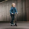 A young boy is riding a black stunt scooter on a paved surface with a textured gray wall in the background. Náhled obrázku produktu