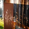 A decorative metal screen with a dandelion motif is placed next to a wooden wall, casting shadows. Produktbild-Vorschau