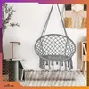 A gray macrame hanging chair with fringe is displayed in a room with a plant, a side table, and a rug. Voir les images du produit