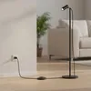 A black floor lamp with a dimmer is plugged into a wall socket in a living room setting. Produktbild-Vorschau