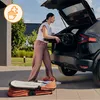 A woman is loading a stroller into the trunk of a car, with a bassinet on the ground nearby. Produktbild-Vorschau