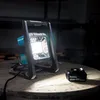 A portable LED work light and a battery are placed on a wooden surface in a dimly lit workshop. Podgląd zdjęcia produktu