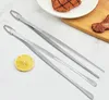 Two stainless steel kitchen tongs are laid out on a marble-like surface, with a slice of lemon and some cooked food in the background. Produktbild-Vorschau