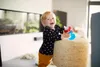 A baby is holding a sippy cup while leaning on a woven ottoman in a living room. Náhled obrázku produktu