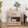 A light wood-colored nightstand with a drawer and an open shelf, placed next to a bed. The top of the nightstand is decorated with a vase of flowers, a clock, and a diffuser. Podgląd zdjęcia produktu