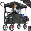 A girl sits in a black folding wagon with a canopy, which is loaded with items. The wagon has four wheels and a handle for pulling. Produktbild-Vorschau