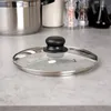 A glass pot lid with a black handle sits on a speckled gray countertop. Náhled obrázku produktu
