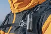 A person wearing a backpack and jacket, with a small cylindrical device attached to the backpack strap. Podgląd zdjęcia produktu