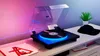 A turntable with LED lighting is placed on a shelf next to a baseball cap and a potted plant. Two record sleeves are in the foreground. Anteprima dell’immagine prodotto