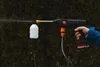 A hand holding a cordless pressure washer with a foam cannon attachment, spraying water. Náhled obrázku produktu
