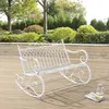 A white metal rocking bench is placed on a wooden deck in a garden setting with lush green bushes and trees in the background. Náhled obrázku produktu