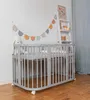 A baby crib with a mattress and bedding, decorated with a colorful garland and a stuffed bunny, placed next to a ladder shelf in a nursery. Produktbild-Vorschau