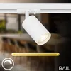 A white track light fixture is mounted on a white track, angled downwards. The background shows a blurred interior scene with furniture. A yellow gradient bar and a white icon with a crescent shape are visible at the bottom. Náhľad obrázku produktu