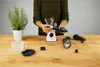 A person's hands assembling a white and black juicer on a wooden table, with a potted plant and other juicer parts nearby. Produktbild-Vorschau