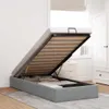 A grey ottoman bed with the mattress lifted, revealing storage space underneath. The bed is in a bedroom setting with a white wardrobe and a chest of drawers visible in the background. Produktbild-Vorschau