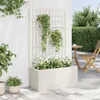 A white planter box with a trellis is shown outdoors against a white wall, with plants growing in and on it. A wooden table and chair are visible in the background. Podgląd zdjęcia produktu