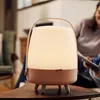 A portable lamp with a Bluetooth speaker sits on a wooden table in a living room setting, with people in the background. Produktbild-Vorschau