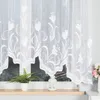 A white lace curtain with a floral pattern hangs over a window. A potted fern and a white lantern sit on the windowsill. Produktbild-Vorschau