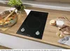 A black electric hob with two burners is shown on a wooden countertop, with a pot of food cooking on one burner. Produktbild-Vorschau