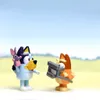 Two Bluey figurines, one holding a pink bunny toy and the other holding a gray camera, stand in a bright, outdoor setting with a blurred background. Voir les images du produit