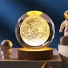 A crystal sphere with a 3D moon inside, illuminated from below, sits on a wooden base. Two small astronaut figurines are placed on either side of the lamp. Náhled obrázku produktu