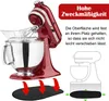 A red stand mixer with a clear bowl and whisk attachment is shown from a diagonal angle. Below the mixer, a black anti-slip mat is depicted with red arrows indicating it should be placed underneath. A smaller, outlined illustration of a mixer on a mat is s Produktbild-Vorschau