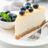 A slice of cheesecake with blueberries and a mint leaf on a white plate, with a fork. Produktbild-Vorschau