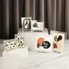 Several framed abstract art pieces are displayed on white pedestals against a dark brown curtain background. Produktbild-Vorschau